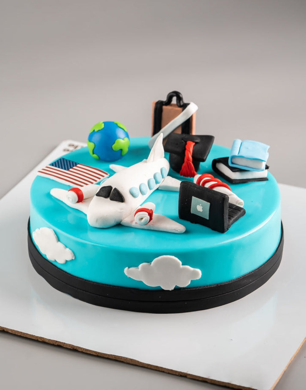 Travel and graduation theme designer cake with airplane, globe, laptop, books, and suitcase decorations, perfect for farewell or study abroad celebration.
