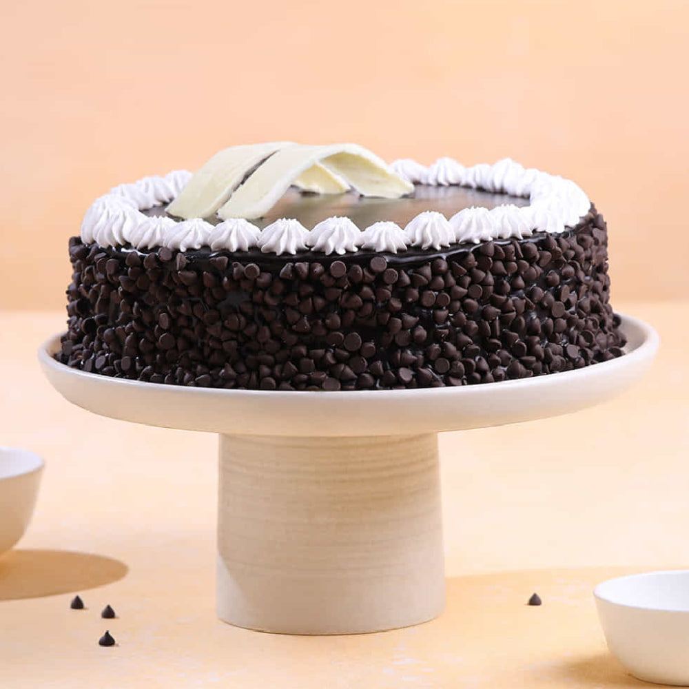 A decadent chocolate cake covered in dark chocolate chips, topped with smooth chocolate ganache, white chocolate curls, and a decorative border of whipped cream frosting on a white cake stand. side view