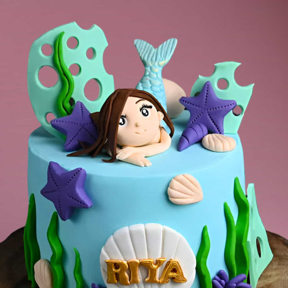 mermaid-themed birthday cake with blue fondant, edible seashells, purple starfish, and a mermaid topper.