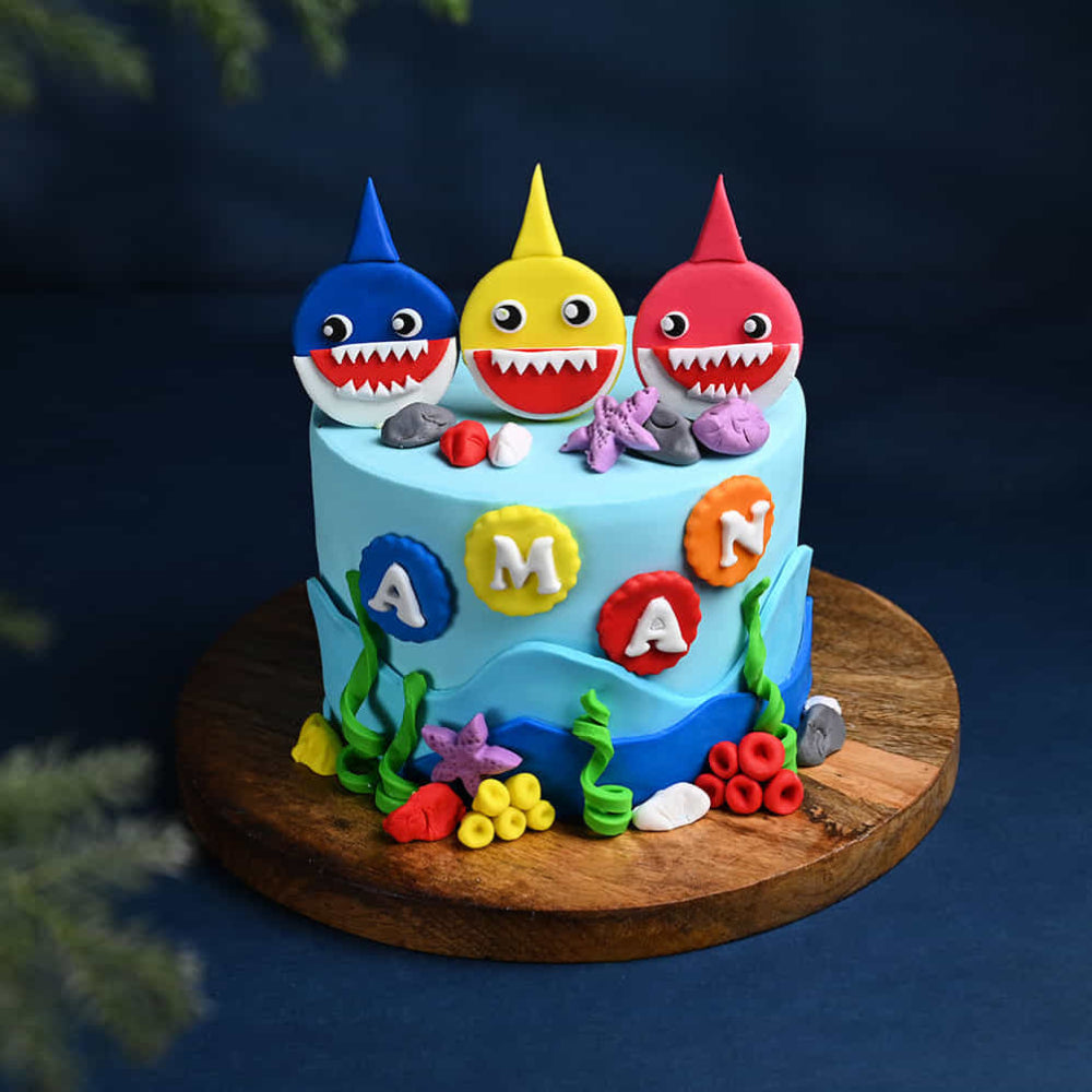 Light blue Baby Shark birthday cake for Aman featuring red, yellow, and blue shark toppers and underwater coral decorations.