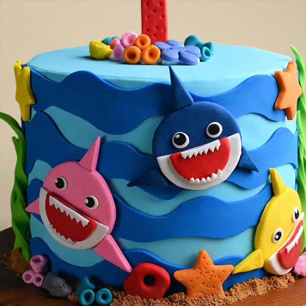 blue Baby Shark birthday cake with yellow, pink, and blue shark characters, coral decorations, and a number 1 topper.
