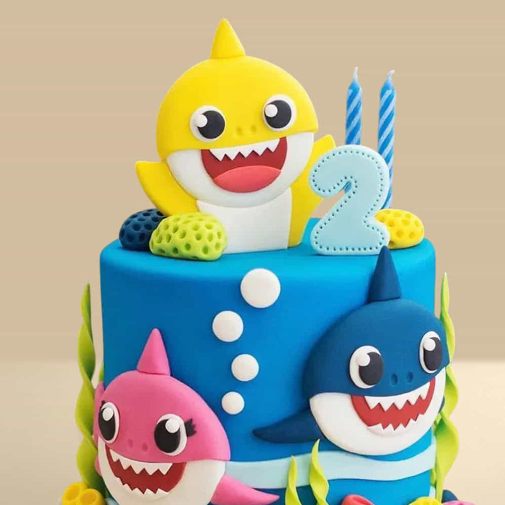 blue Baby Shark birthday cake with yellow, pink, and blue shark characters, coral decorations, and a number 2 topper.