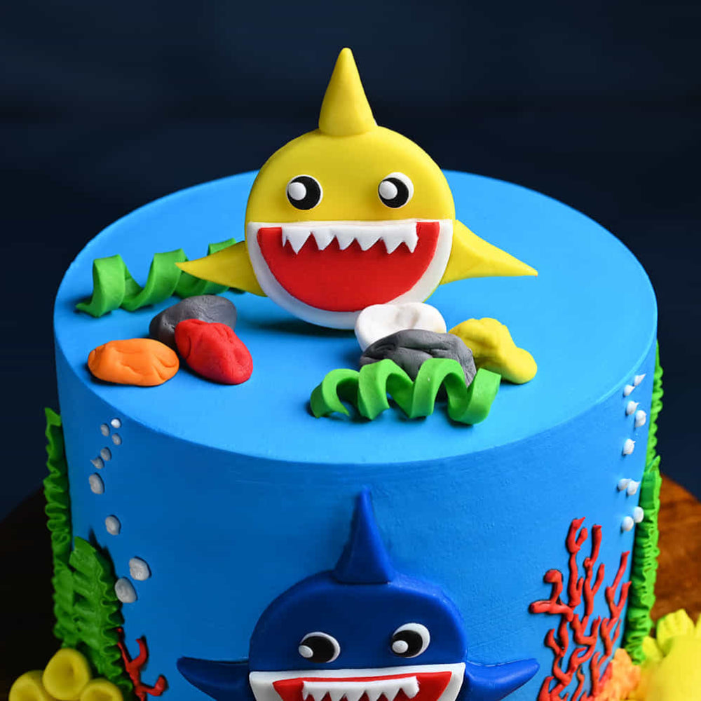 Blue ocean-themed Baby Shark cake with 3D yellow and blue shark toppers, edible coral, and sea plants.