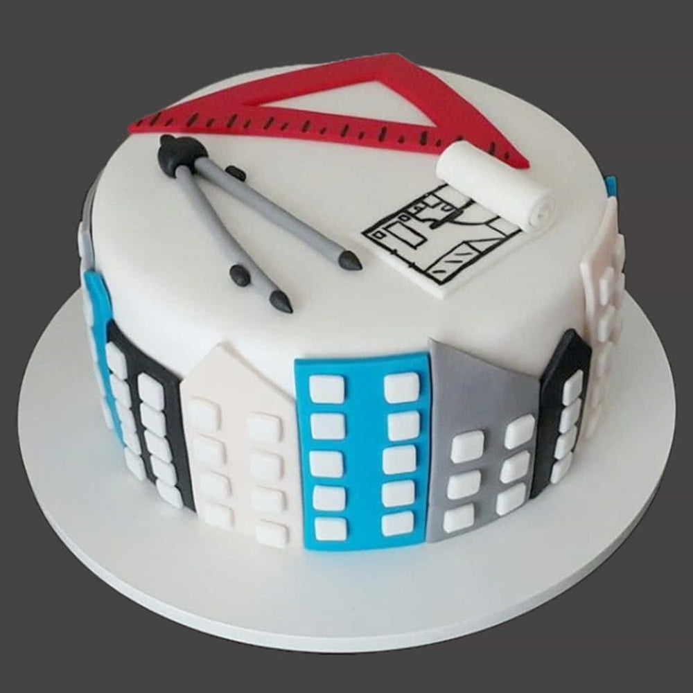 White round cake decorated with an architecture theme, featuring fondant tools like a triangular ruler, compass, and blueprint on top, with building designs around the sides.