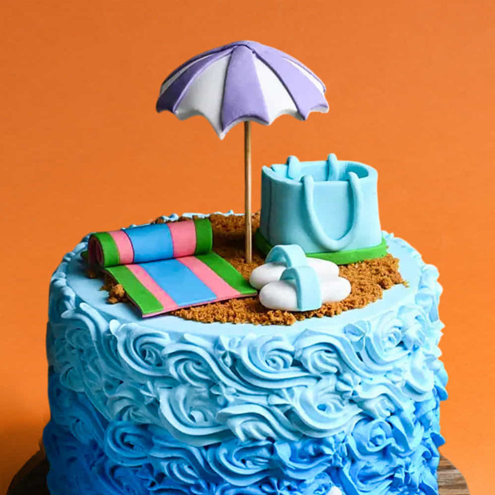 Beach theme designer cake with umbrella topper, towel, sand, and bag decorations, featuring blue wave design, perfect for summer or vacation celebration.