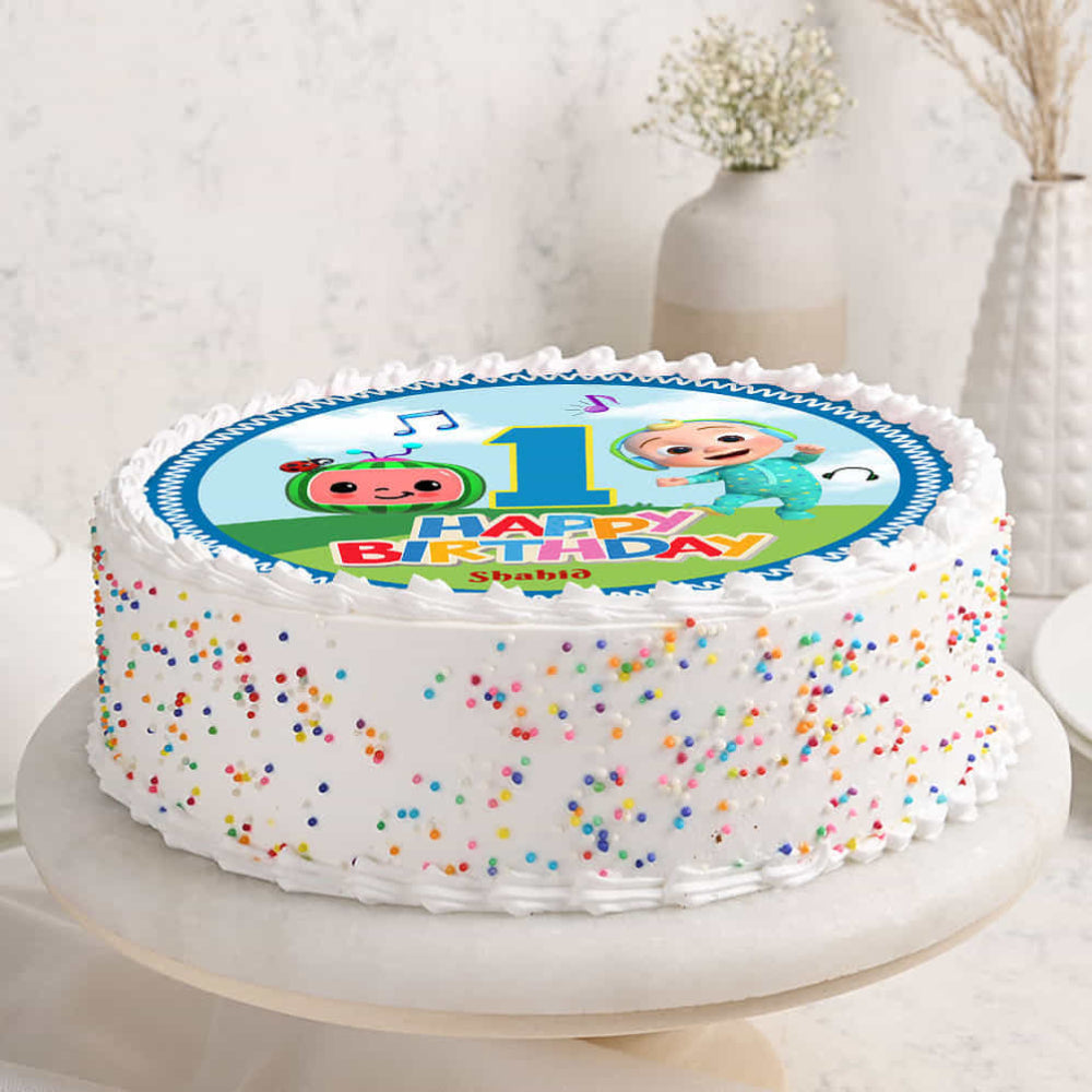 A round 1st birthday cake with white frosting and colorful sprinkles on the sides. The top features a Cocomelon edible image with JJ, the Cocomelon logo, a large number 1, and the name "Shahid.