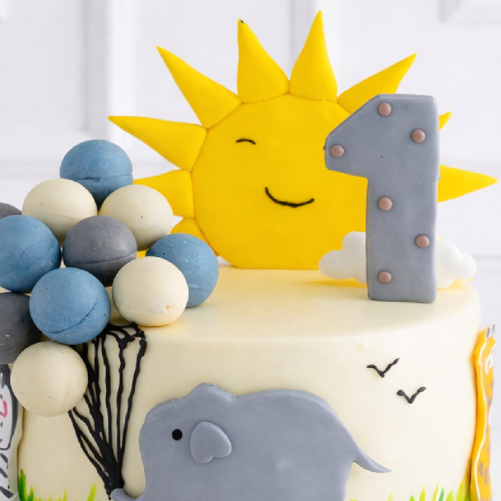 Cute first birthday designer cake with number one topper, smiling sun decoration, pastel balls, and elephant theme, perfect for a baby celebration.