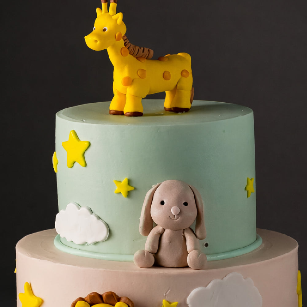 Two-tier pastel designer cake with fondant giraffe topper, teddy bunny figure, stars, and clouds, perfect for a cute kids birthday celebration.