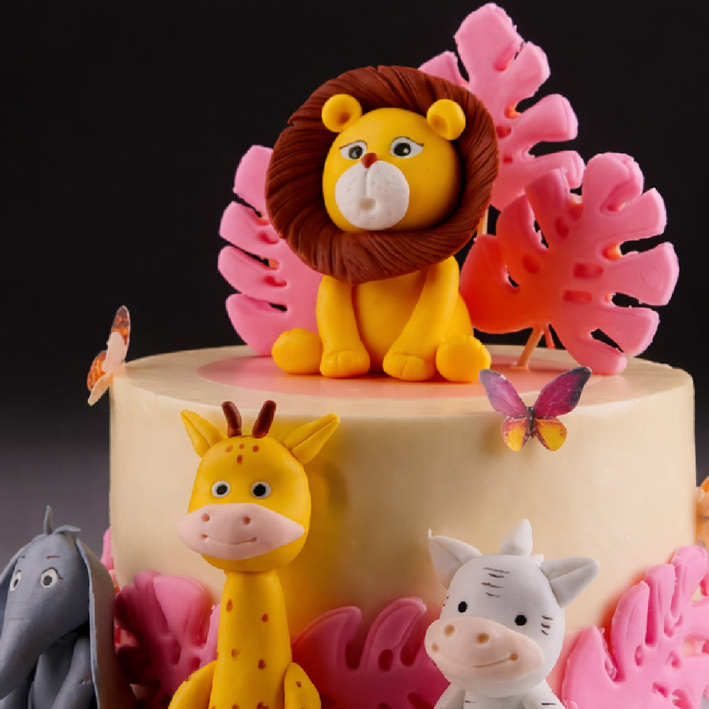 Cute jungle theme designer cake with lion topper, giraffe, zebra, and elephant figures, decorated with pink leaves and butterflies, perfect for a kids birthday celebration.
