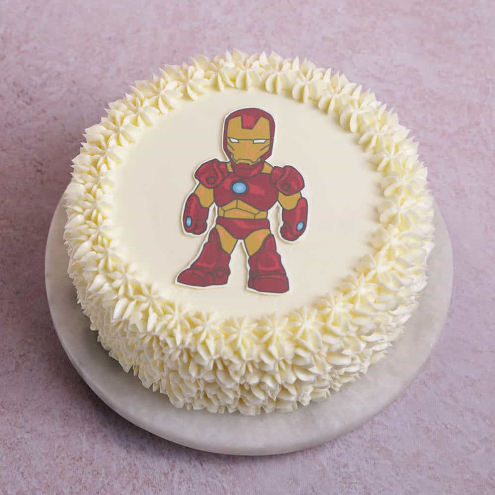 Round cake covered in white frosting with piped star decorations around the edges, featuring a cartoon-style red-and-gold armored superhero figure printed on the center. The cake sits on a white board against a light pink background.