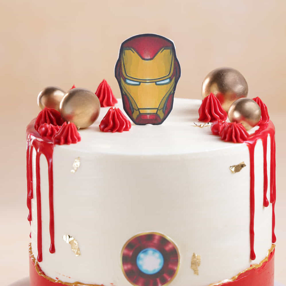 White frosted round cake with red drip icing, topped with gold decorative spheres, red dollops of frosting, and a cutout of a red-and-gold superhero helmet. The cake also features small gold accents and a circular emblem on the side, set against a neutral background.