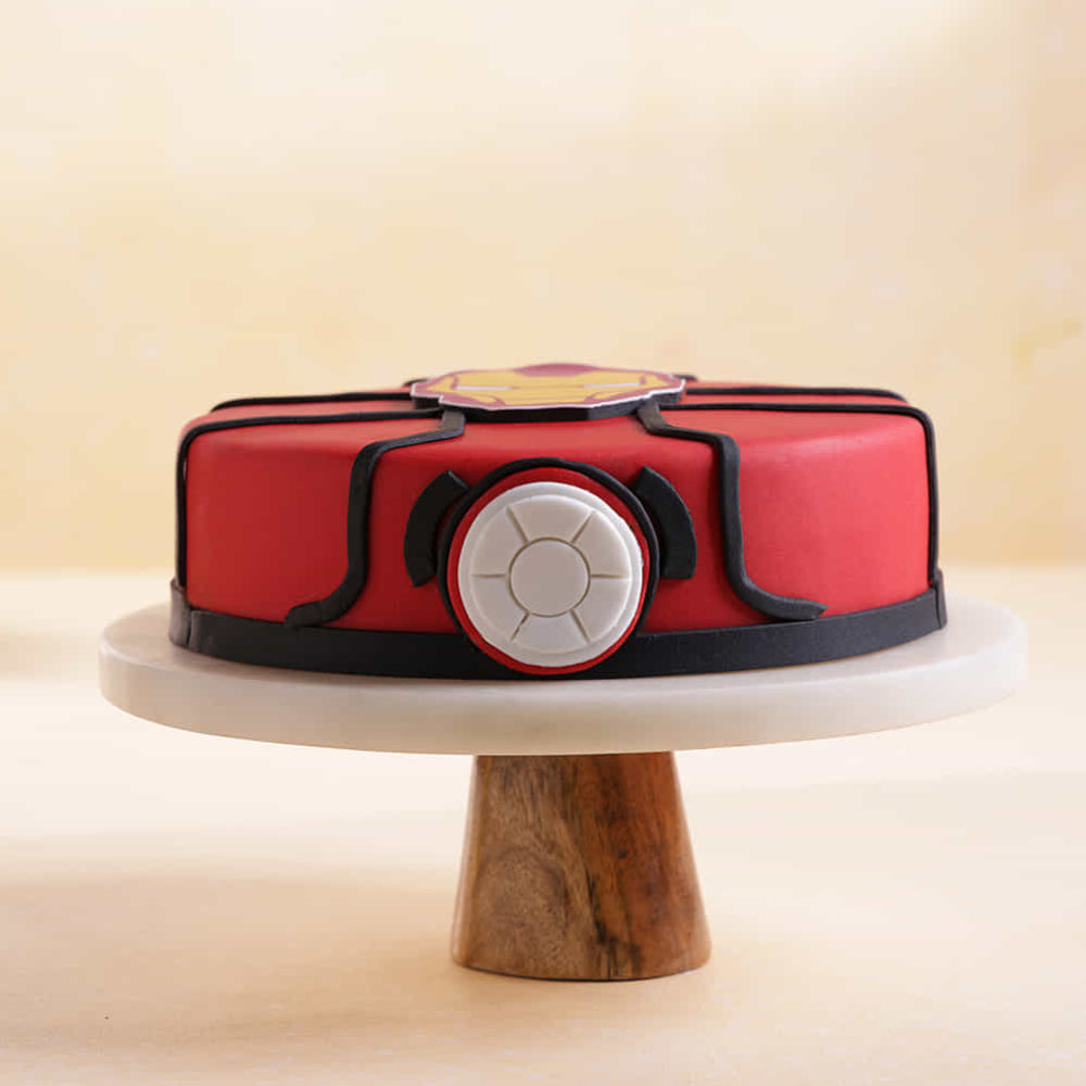 Red round cake on a pedestal stand, decorated with black fondant lines and a central circular emblem resembling a superhero suit design, set against a soft beige background.