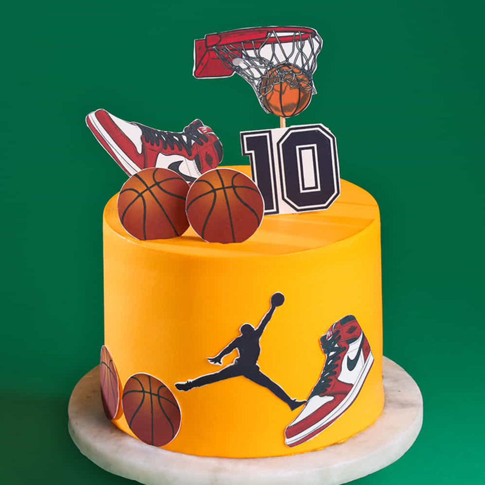 A tall, vibrant yellow birthday cake featuring Air Jordan sneakers and basketball decorations. The cake is topped with a basketball hoop, a number "10" sign, and basketball cutouts, with a Jumpman silhouette and sneaker images on the side.