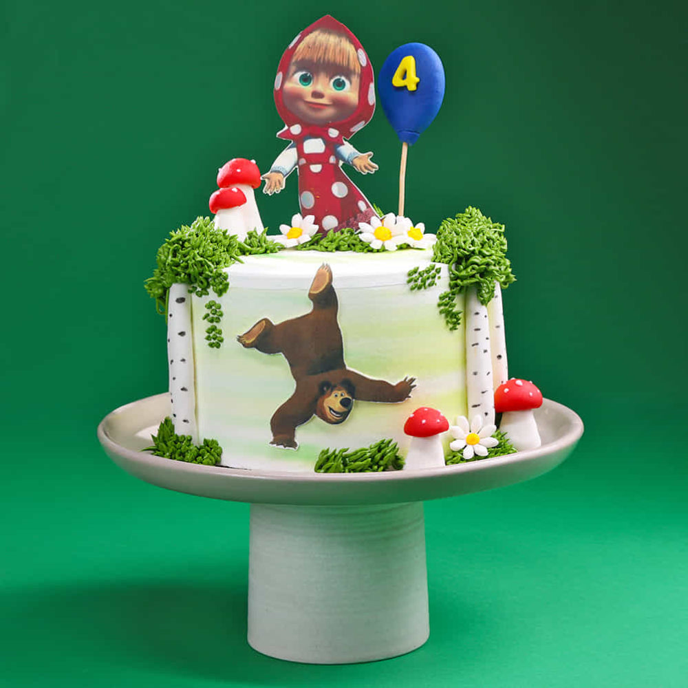 White round cake decorated with a forest theme, featuring green grass-like frosting, small red mushrooms, and flowers. On top is a cartoon girl in a red hood holding a blue balloon with the number “4,” and the side of the cake shows a playful bear illustration. The cake is displayed on a pedestal against a green background.