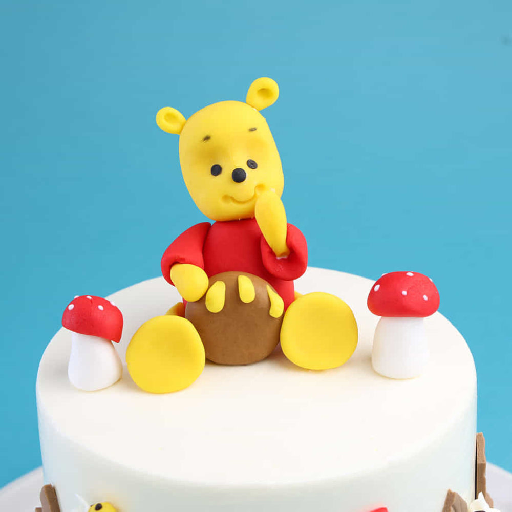White frosted cake topped with a fondant figure of a yellow bear in a red shirt sitting with one hand to its mouth. The cake is decorated with small red-and-white mushroom accents and set against a blue background.