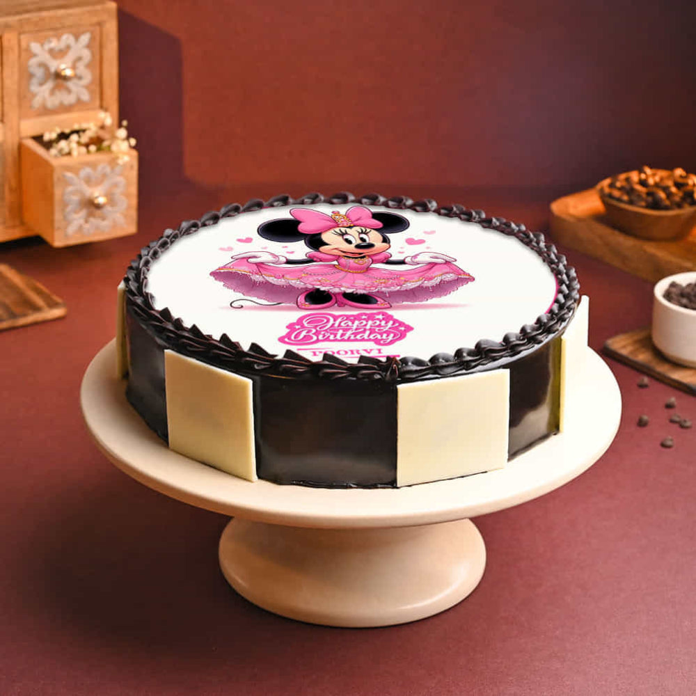 Round cake with glossy dark chocolate sides and a piped chocolate border, featuring an edible image of a cartoon mouse character in a pink dress on top with a “Happy Birthday” message. The cake is displayed on a pedestal stand against a warm-toned background with decorative items nearby.