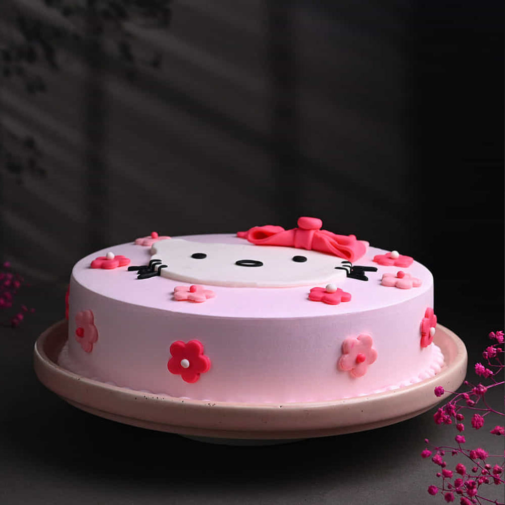 Pastel pink round cake with a cute cartoon-style face on top, decorated with a red bow and small pink flowers around the sides. The cake sits on a plate with a dark, softly lit background and delicate floral accents nearby.
