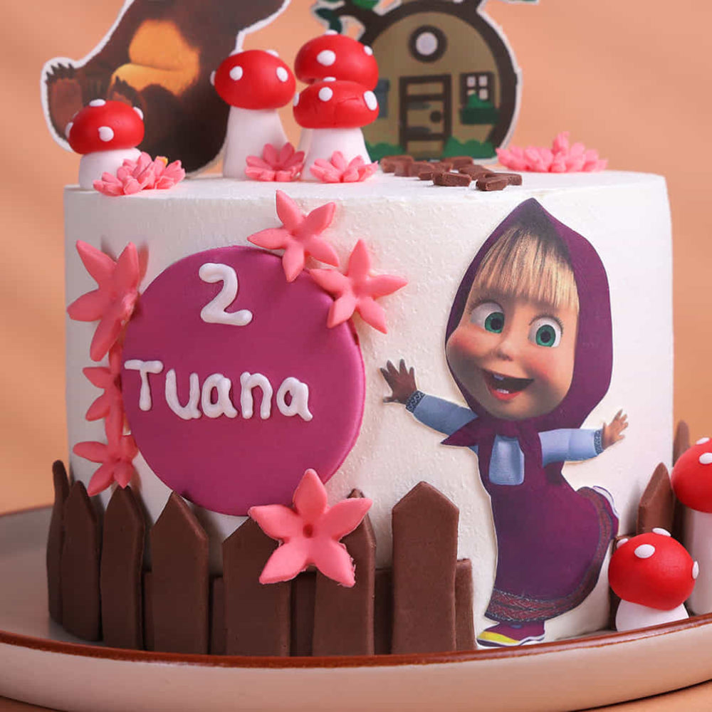 Close-up of a themed cake decorated White round cake decorated with a woodland theme, featuring red mushrooms, pink flowers, and a small fence design around the base. The cake includes cartoon cutouts of a girl in a hood and a bear, along with a house topper, and a pink plaque that reads “2 Tuana.” It is displayed on a plate against a warm beige background.