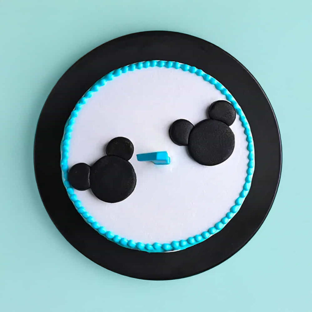 Minimalist round cake with smooth white frosting, decorated with a turquoise border and black fondant shapes resembling two cartoon mouse heads on top. The cake sits on a black plate against a light blue background.