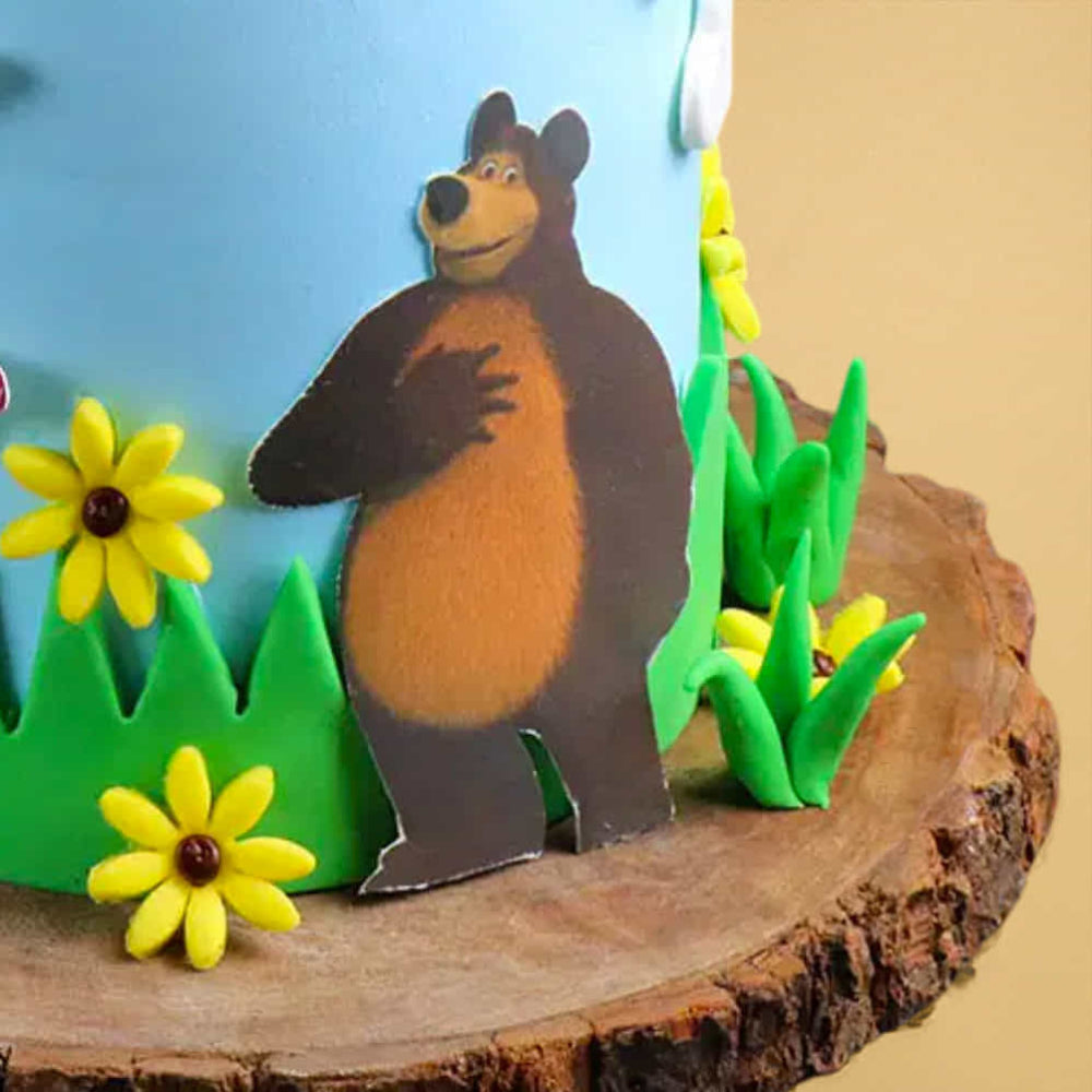 Close-up of a themed cake decorated with a cartoon bear cutout standing among green fondant grass and yellow flowers. The cake sits on a wooden-style base, with part of the blue-frosted cake visible in the background.