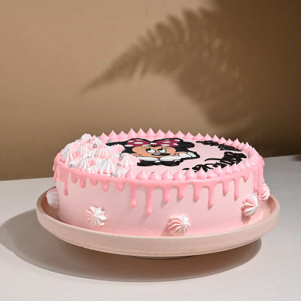 Pastel pink round cake with drip icing, decorated with white whipped frosting swirls and small dollops along the sides. The top features a cartoon mouse character design, and the cake is placed on a plate against a warm, softly lit background with a plant shadow.