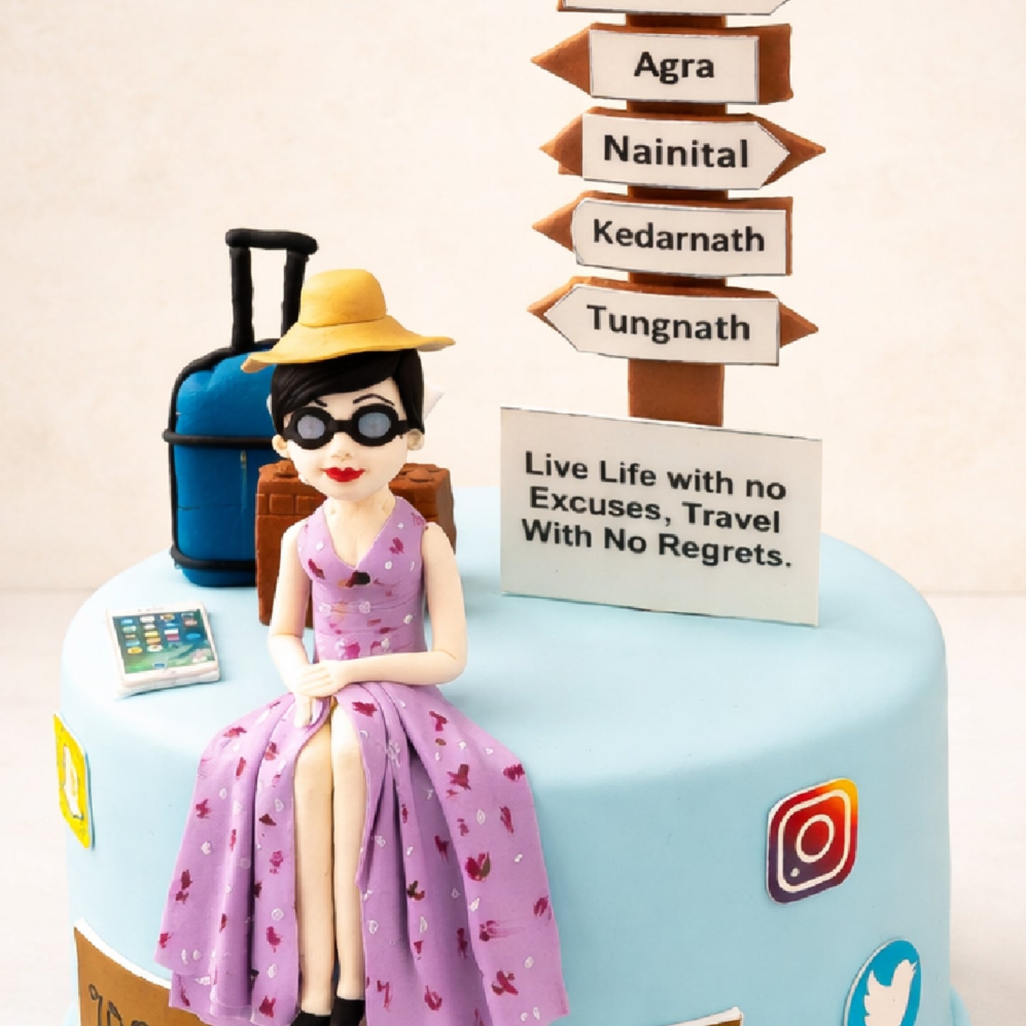 Travel-themed cake with a seated fondant figure of a woman wearing a hat and sunglasses, next to a suitcase and a signpost listing destinations. The cake features a message reading “Live Life with no Excuses, Travel With No Regrets.” and travel-related decorations.