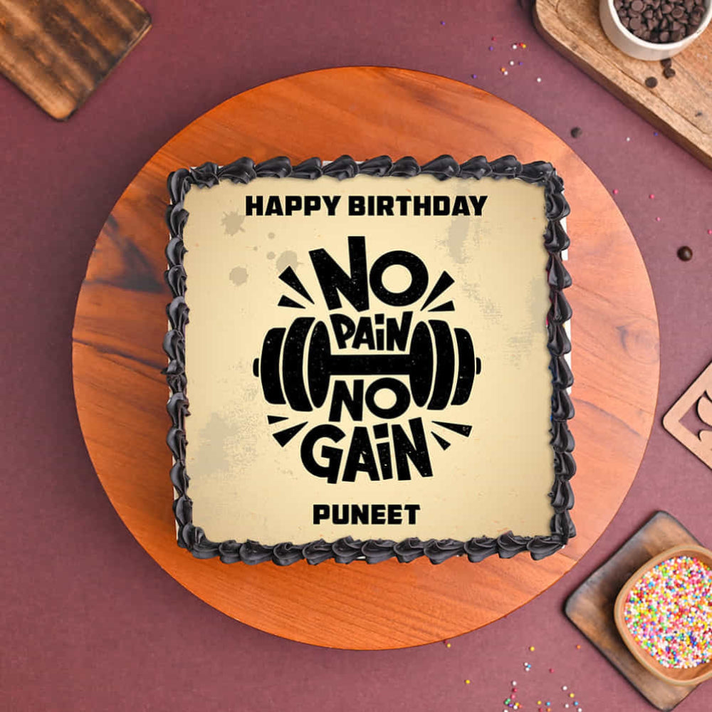 Square cake with a black frosting border and a printed message reading “Happy Birthday No Pain No Gain Puneet,” featuring a barbell graphic, placed on a wooden board.