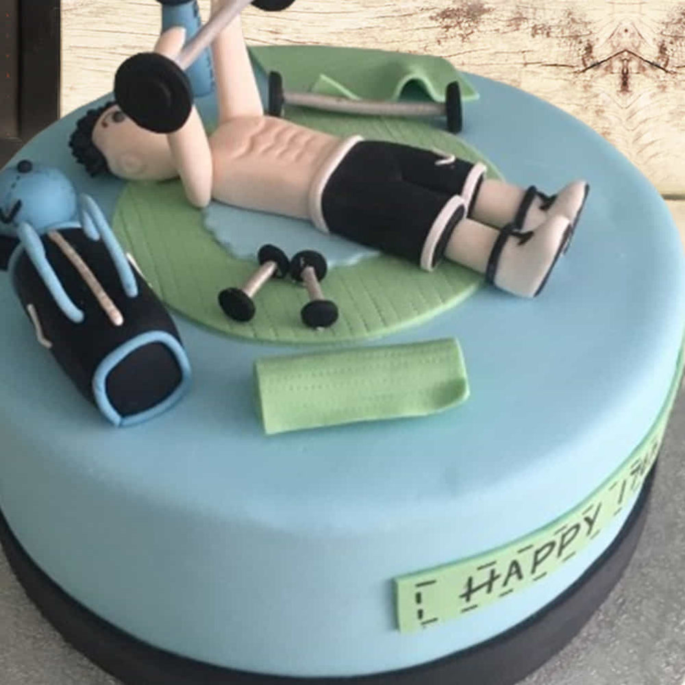 Blue round cake with a gym theme, featuring a fondant figure lifting weights while lying on a bench, surrounded by small dumbbells, a gym bag, and workout equipment.