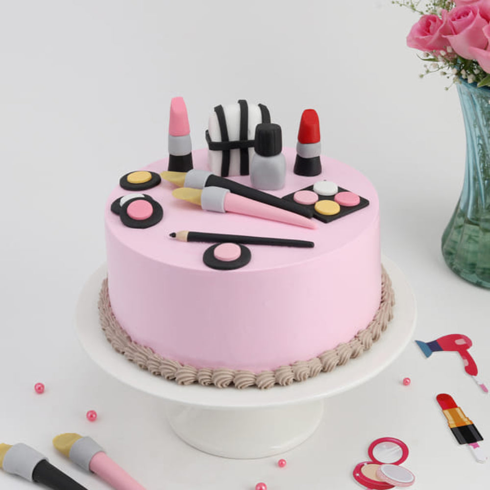 Pink designer cake decorated with makeup items like lipstick, nail polish, brushes, and cosmetics, placed on a cake stand with a clean background and floral vase.