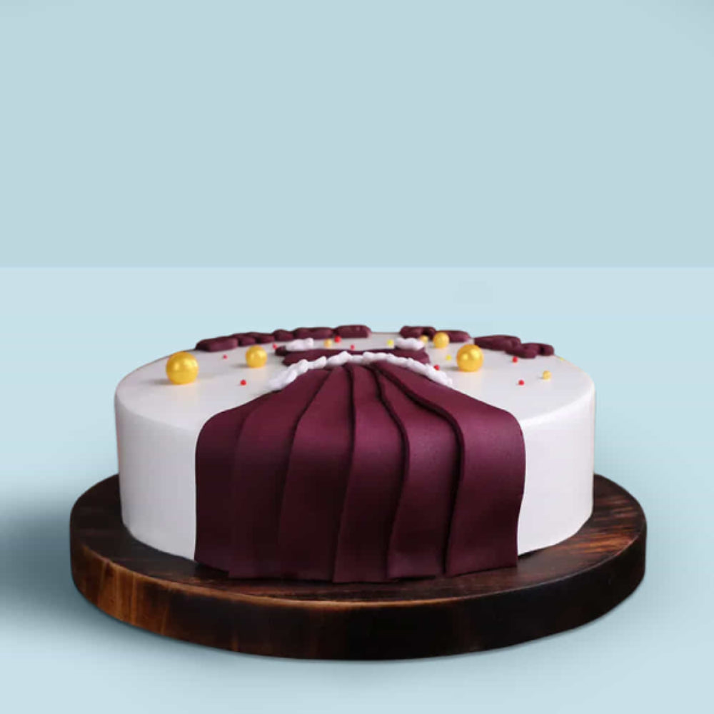 Elegant white designer cake with maroon fondant drape and gold decorations on a wooden base, perfect for special occasions in Delhi.