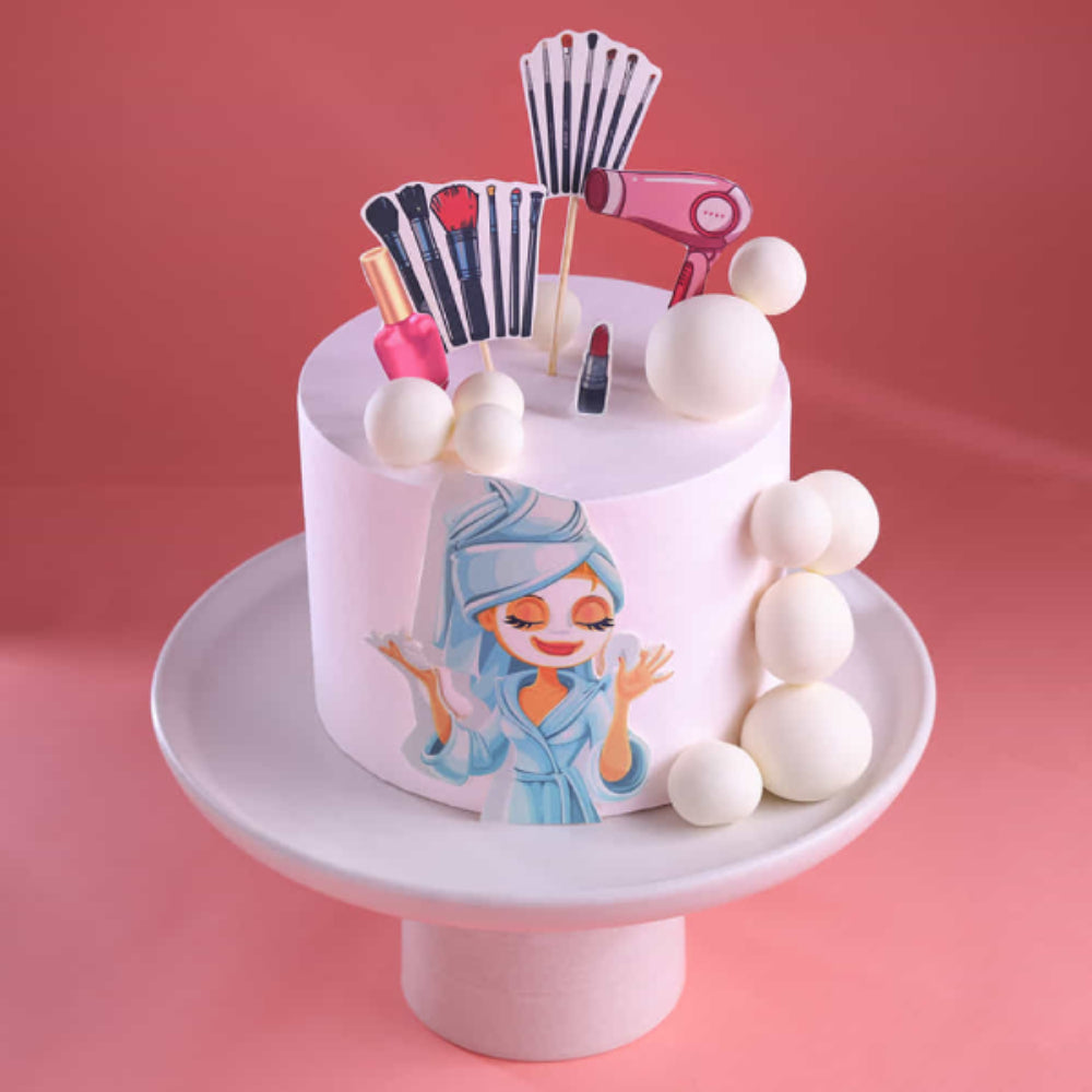 Beauty-themed designer cake with makeup tools like brushes, nail polish, lipstick, and hair dryer, featuring a cartoon girl illustration on a white cake with decorative spheres on a cake stand.
