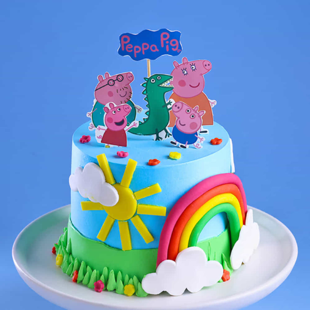 A vibrant two-tier themed cake for kids featuring Peppa Pig characters, a colorful rainbow, and a bright yellow sun. The cake is decorated with light blue and green frosting to resemble a sunny day in a field.