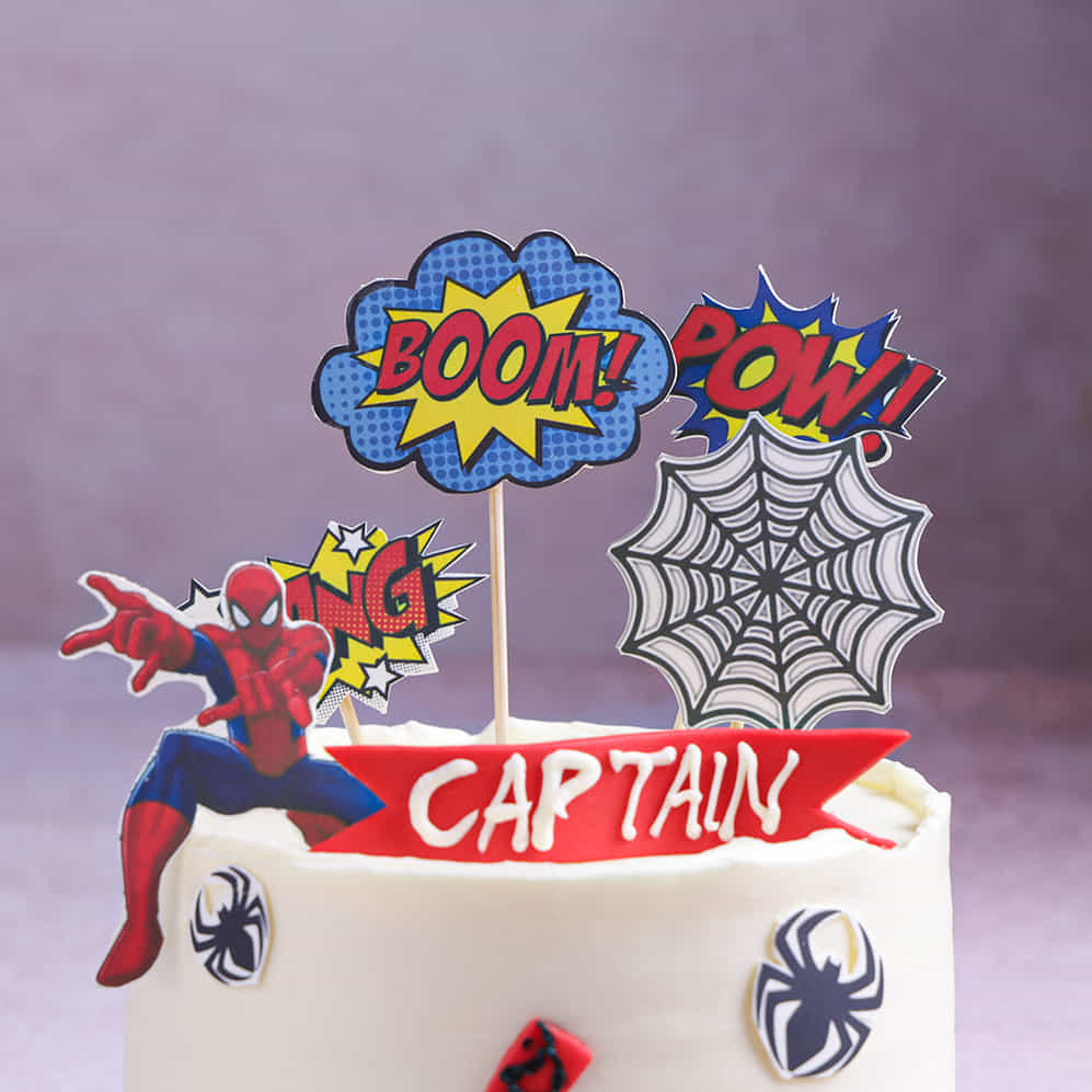Superhero-themed cake with Spider-Man figure and 'BOOM!' 'POW!' toppers on a purple background