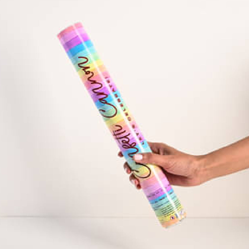 A hand holding a colorful cylindrical confetti popper with rainbow stripes and decorative text, set against a plain light background.