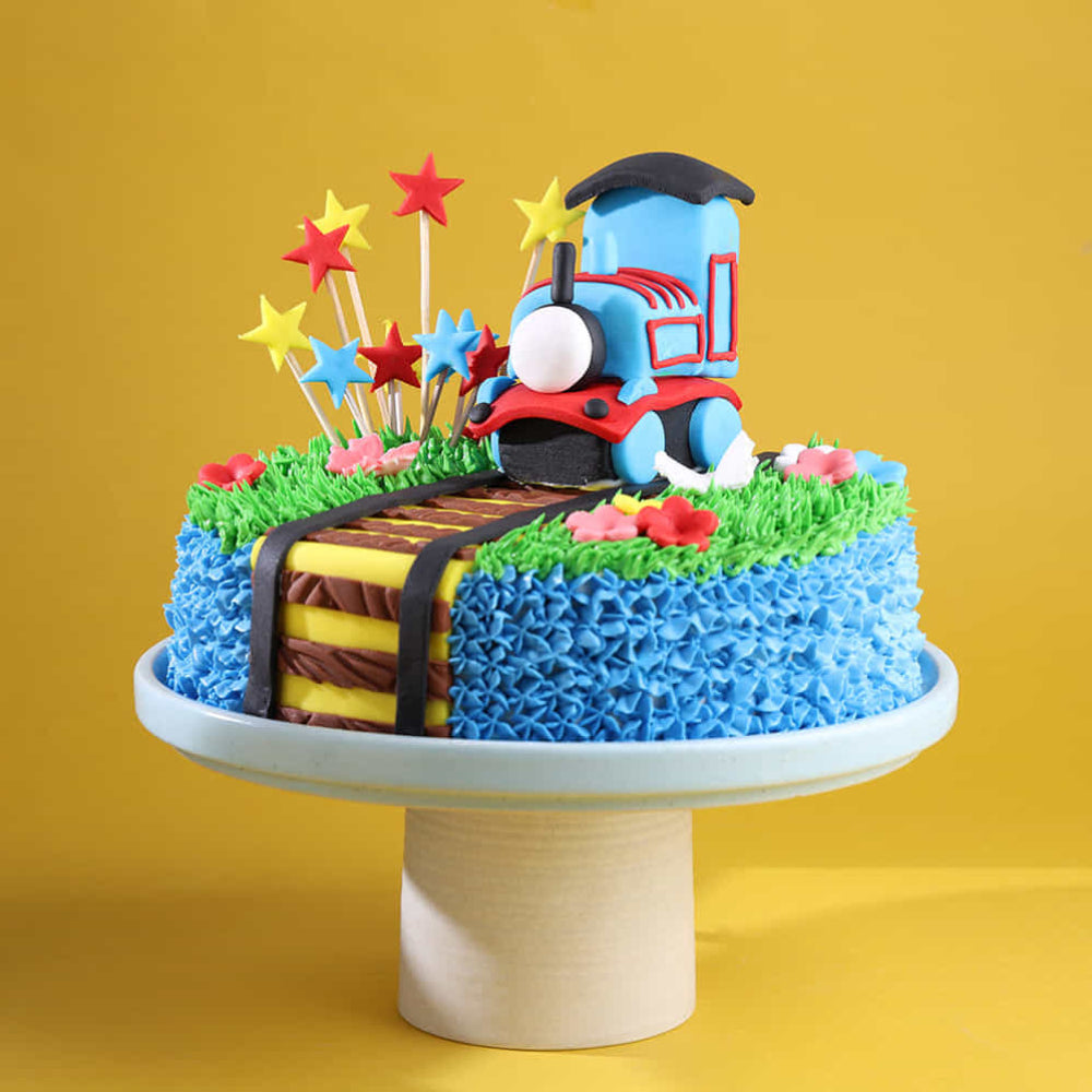 Blue train engine birthday cake with 3D fondant locomotive, railway tracks, and star-shaped cake toppers.