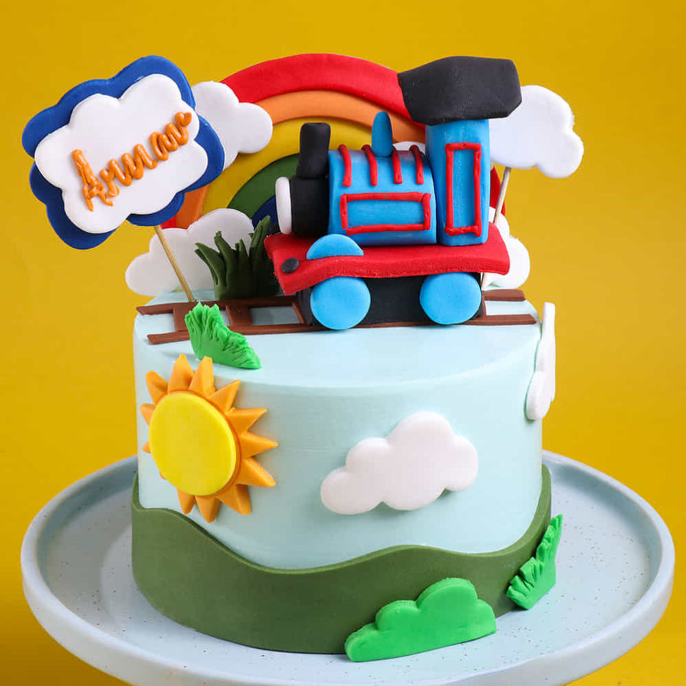 A colorful round cake with light blue frosting on a plate, decorated with a fondant toy train on top sitting on tracks beneath a rainbow. The cake features clouds, a bright yellow sun, green hills, and a sign with a name written on it, all set against a yellow background.
