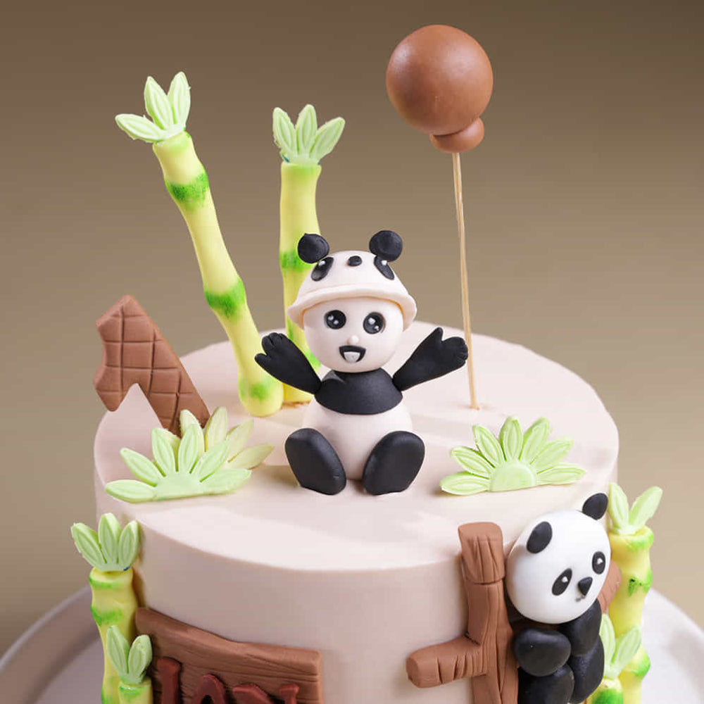A cream-colored first birthday cake featuring three panda figurines and green bamboo stalks.
