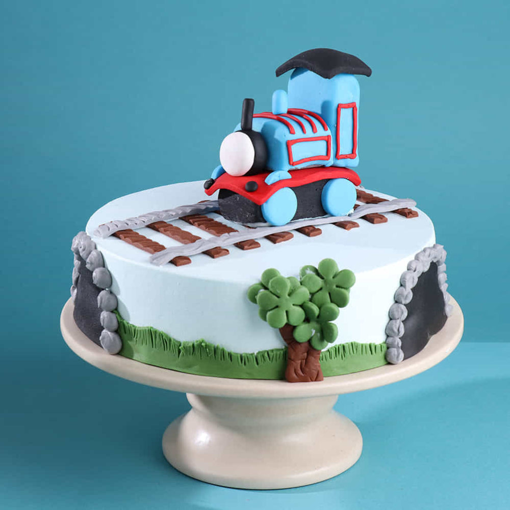 A light blue round cake on a pedestal stand, decorated with a fondant toy train on top sitting on tracks. The sides feature tunnel designs, green grass, and a small tree, all set against a blue background.
