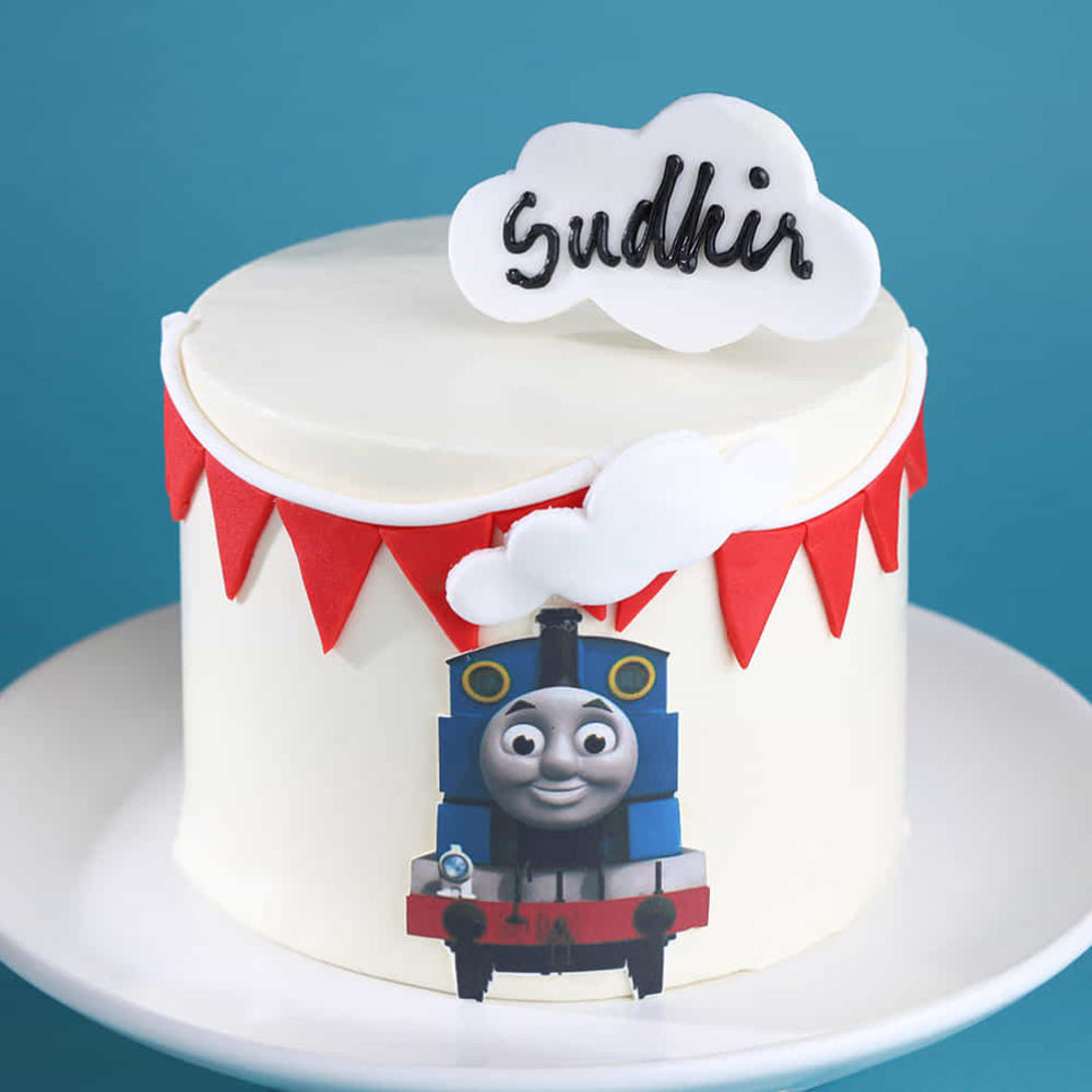 A white frosted round cake on a pedestal stand against a blue background, decorated with red bunting and a smiling cartoon train on the front. A cloud-shaped topper on