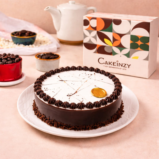 A round chocolate truffle cake with a smooth white glaze top, dark chocolate piped borders, and a gold chocolate coin accent, displayed with a Cakeinzy brand box.