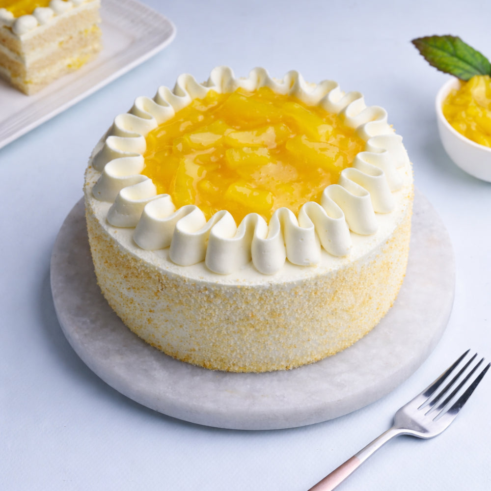 A round vanilla sponge cake coated in fine cake crumbs, topped with a wavy ring of white whipped cream frosting and a bright yellow pineapple glaze filling.