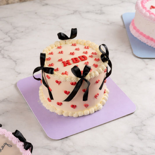 A minimalist cream birthday cake decorated with small red hearts, black ribbon bows, and 'HBD' lettering on a purple base.