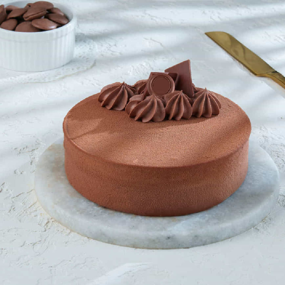A gourmet, minimalist dark chocolate mousse cake served on a marble platter with chocolate shards and ganache swirls on top, accompanied by a small bowl of chocolate discs and a gold knife. top angle view