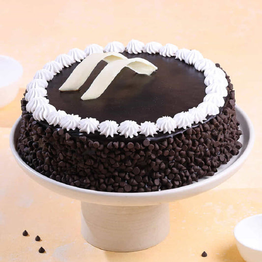 A decadent chocolate cake covered in dark chocolate chips, topped with smooth chocolate ganache, white chocolate curls, and a decorative border of whipped cream frosting on a white cake stand.