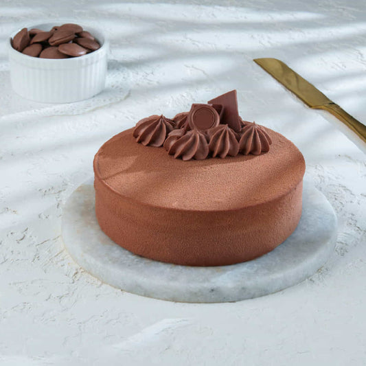 A gourmet, minimalist dark chocolate mousse cake served on a marble platter with chocolate shards and ganache swirls on top, accompanied by a small bowl of chocolate discs and a gold knife.