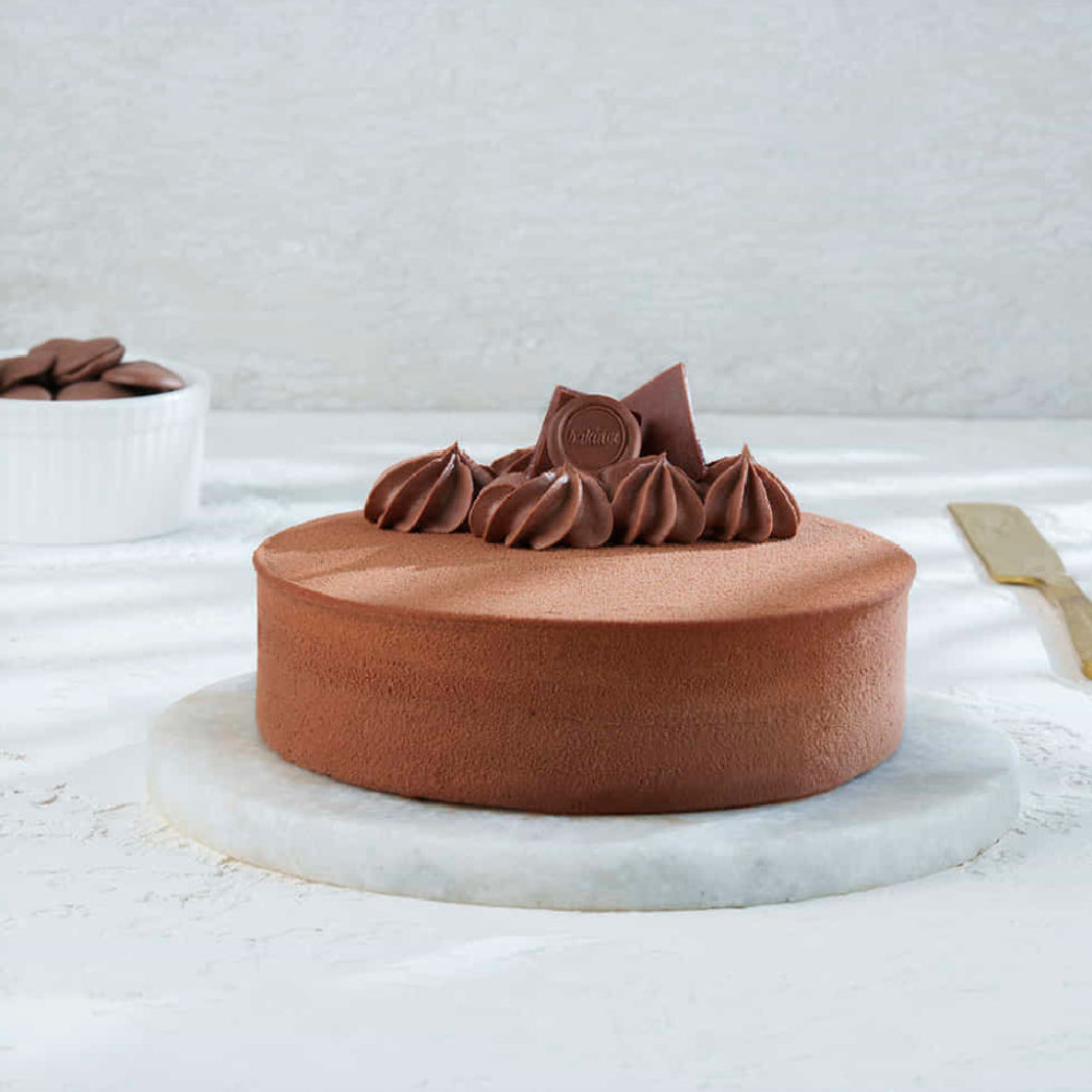 A gourmet, minimalist dark chocolate mousse cake served on a marble platter with chocolate shards and ganache swirls on top, accompanied by a small bowl of chocolate discs and a gold knife. side view