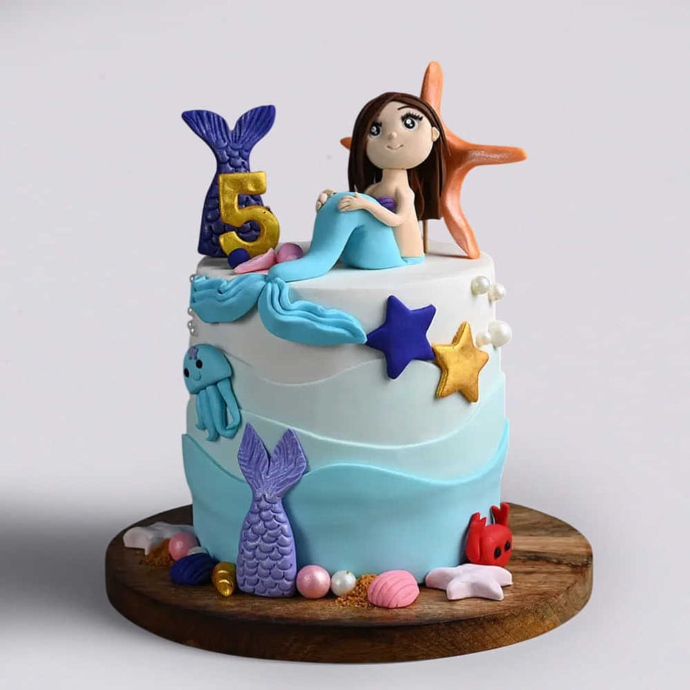 Two-tier blue and white mermaid birthday cake for a 5-year-old featuring a mermaid topper, starfish, and gold "5".