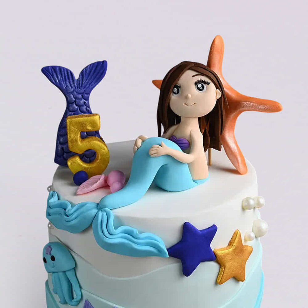 Two-tier blue and white mermaid birthday cake for a 5-year-old featuring a mermaid topper, starfish, and gold "5".