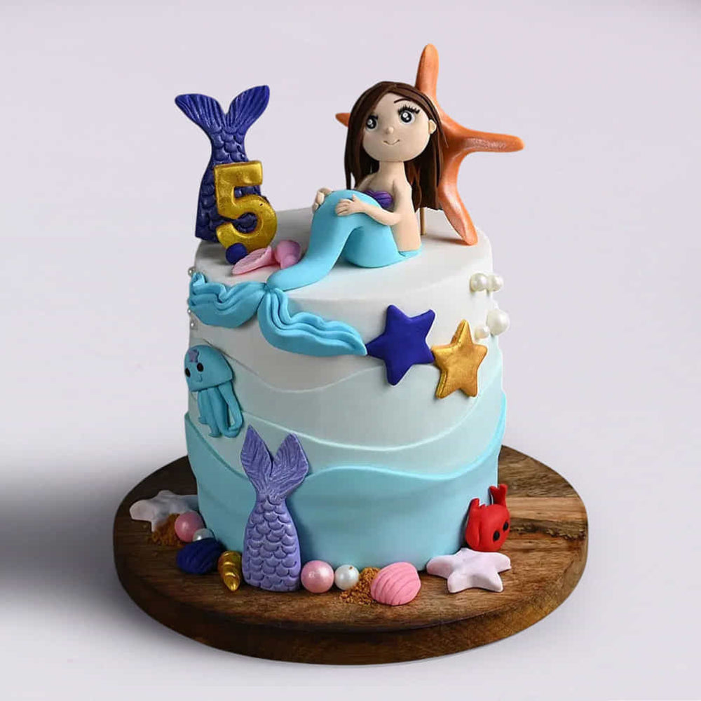 Two-tier blue and white mermaid birthday cake for a 5-year-old featuring a mermaid topper, starfish, and gold "5".