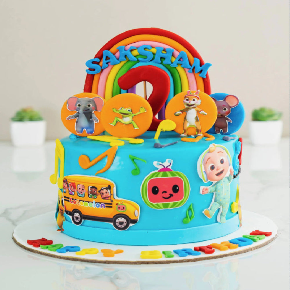 Cocomelon themed designer cake with rainbow topper, number two, cartoon characters, musical notes, and name Saksham, perfect for a kids birthday celebration.